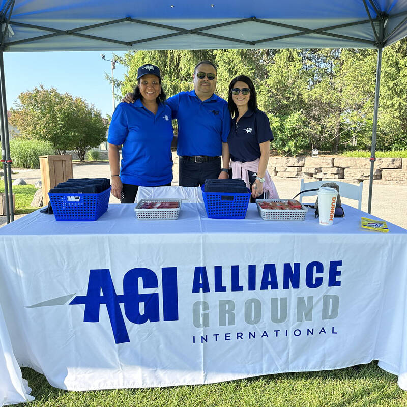 Three individuals stand behind a table covered with a white tablecloth displaying the "AGI Alliance Ground International" logo. The table, set up for the 2023 Golf Outing, has two blue baskets, candies, and promotional pamphlets. They are outdoors under a canopy on a sunny day.