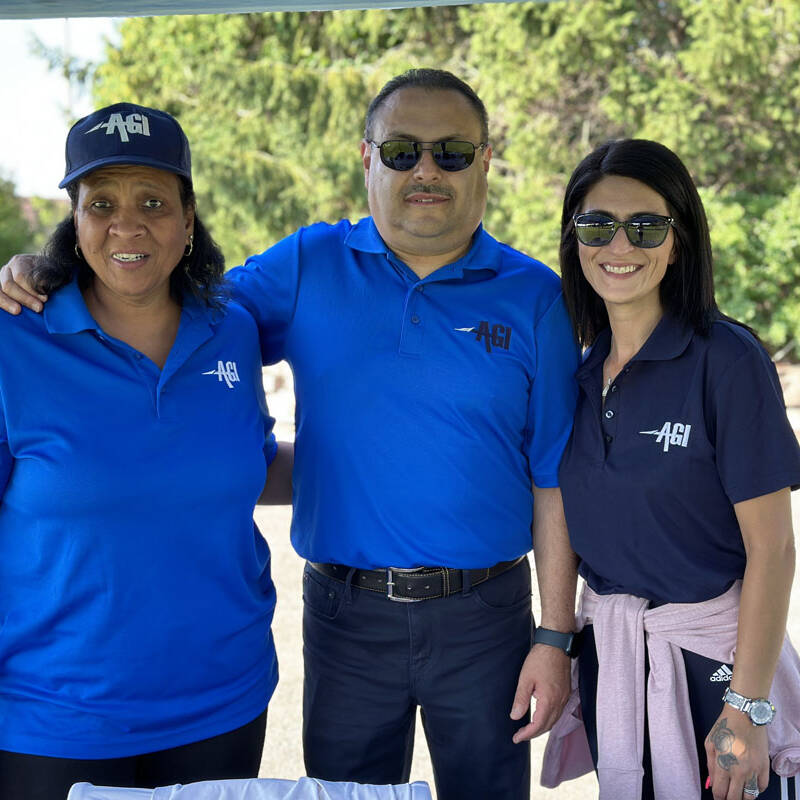 Three people stand outdoors under a shaded area, enjoying a sunny 2023 outing. They are all smiling and wearing matching blue polo shirts with a logo on the left side, perfect for their golf day. The background features lush greenery, and two of them are sporting stylish sunglasses.