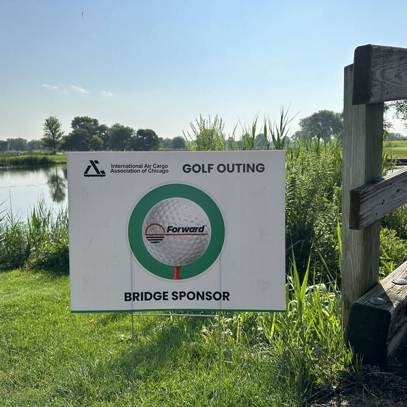 A sign on a grassy area by a lake reads "2023 Golf Outing" with a logo and "International Air Cargo Association of Chicago." "Forward" is printed on the golf ball image inside it. The text at the bottom reads "Bridge Sponsor." A wooden rail is on the right.