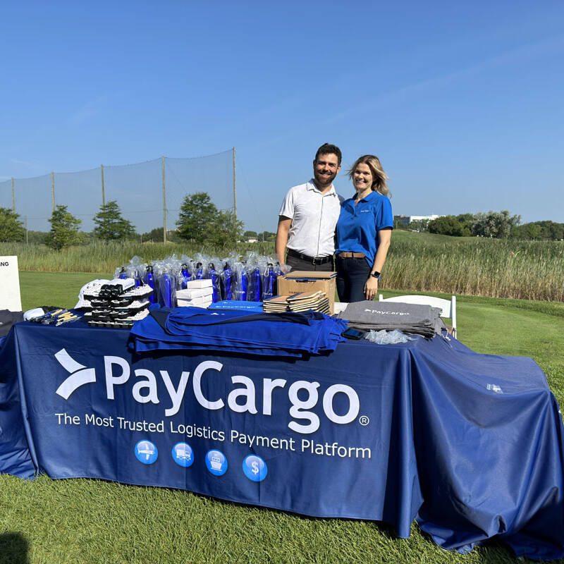 Two people stand behind a 2023 PayCargo-branded table at an outdoor event. The table features various promotional items, including water bottles, bags, and shirts. Behind them, a golf course and a "Golf Outing" sign are visible under the clear blue sky.