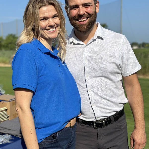 A woman in a blue polo shirt and jeans, and a man in a white patterned shirt and dark pants, stand smiling outdoors on a sunny day at their 2023 golf outing, with a backdrop of greenery and a netted fence.