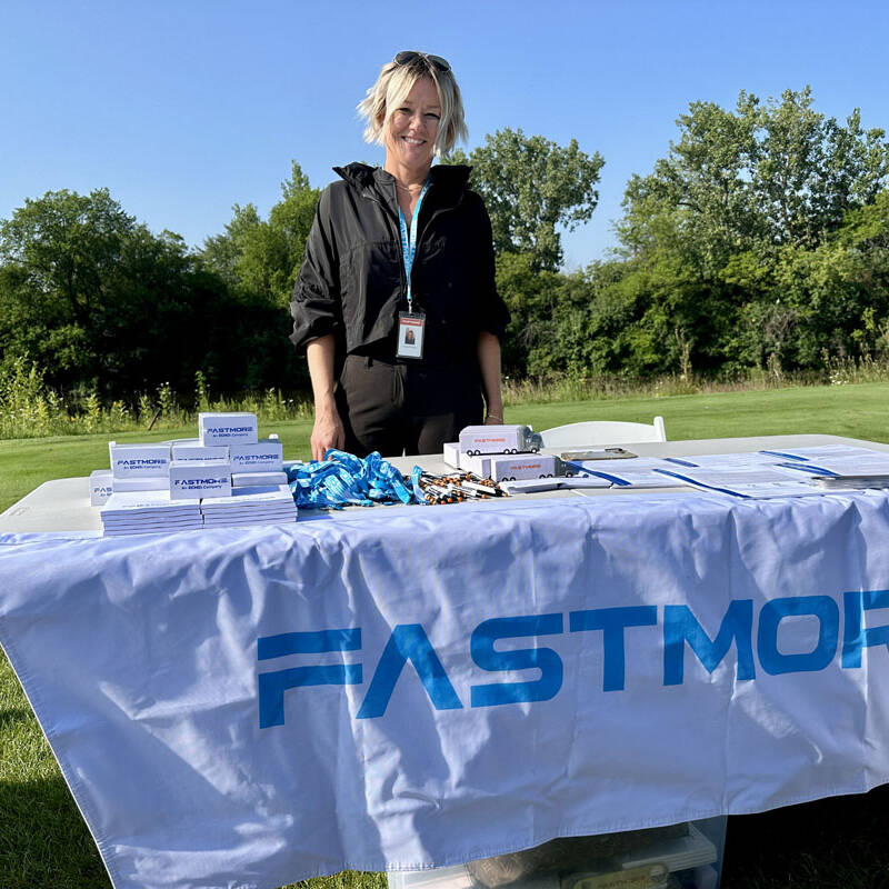 A person wearing a black jacket stands behind a table covered with a white tablecloth printed with the text "FASTMORE." The table holds stacks of small white boxes, lanyards, name tags, and flyers. The backdrop features a lush, green outdoor setting with trees and grass, hinting at a 2023 outing.