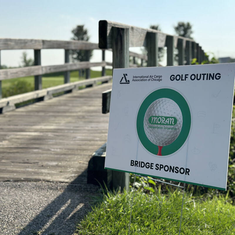 A signboard near a wooden bridge in a green, outdoor setting reads "International Air Cargo Association of Chicago," "Golf Outing 2023," and "Bridge Sponsor." A large golf ball is pictured on the sign with the name "Moran Logistics Company" written on it.