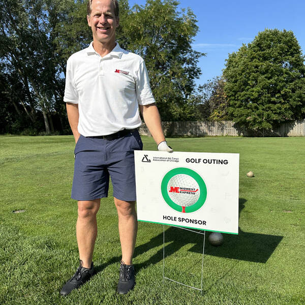 A man in a white polo shirt and navy shorts stands on a grassy golf course next to a sign that reads "GOLF OUTING 2023" and "HOLE SPONSOR." Trees and a wooden fence can be seen in the background, under the sunny weather.