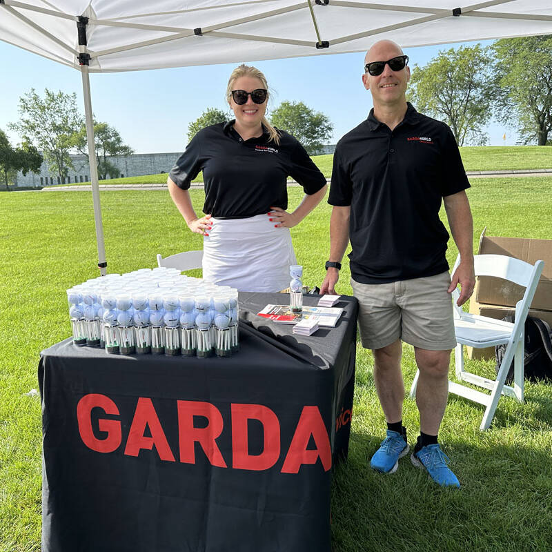 A woman and a man stand under a white canopy tent at an outdoor event, likely the 2023 Golf Outing. Both wear black shirts emblazoned with "GARDA." The table in front of them displays water bottles, lanyards, and pamphlets. The scene is set on a sunny day with green grass and trees around them.