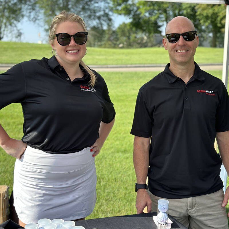 Two individuals, one woman and one man, are standing outside under a canopy during an outing in 2023. Both are wearing black polo shirts with red and white text and sunglasses. The woman has her hand on her hip, and they both are smiling. A grassy lawn and trees, perfect for golf, are visible in the background.