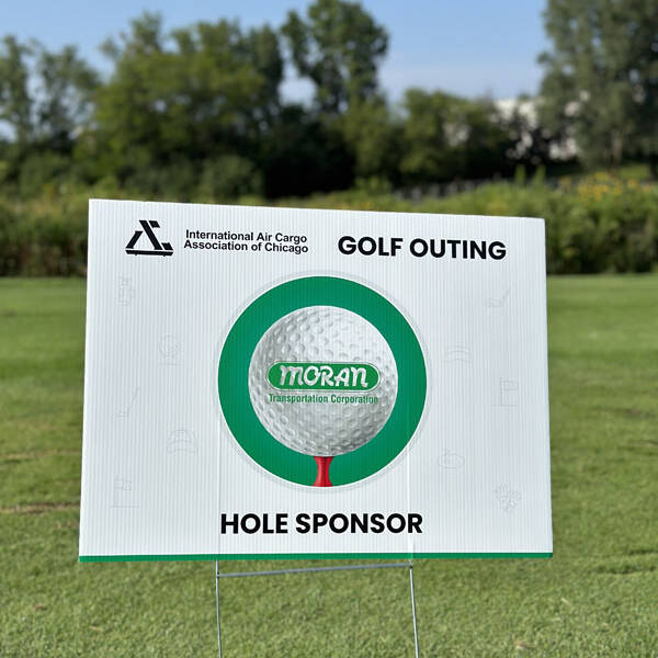 A sign placed on a golf course reads "International Air Cargo Association of Chicago - Golf Outing 2023 - Moran Transportation Corporation - Hole Sponsor." The background displays green grass and trees under clear skies.