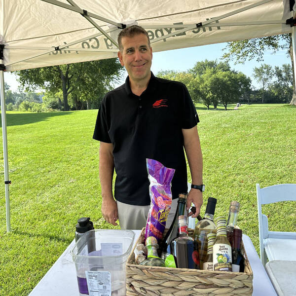 A man in a black polo shirt stands under a canopy tent, smiling. In front of him is a table with a wicker basket containing various bottles and snacks. The background shows a grassy park with trees on a sunny day, perfect for enjoying the 2023 golf outing.