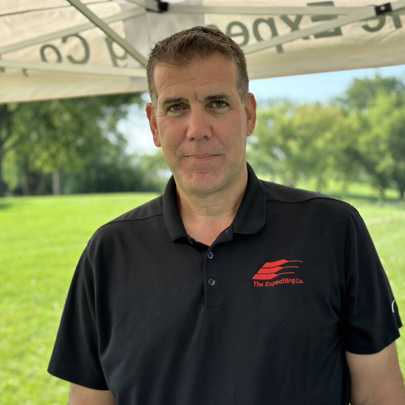 A man stands under a tent outdoors during a sunny golf outing. He is wearing a black polo shirt with a red logo on the chest. The background features green grass and trees, perfectly capturing the essence of 2023's best outdoor activities.