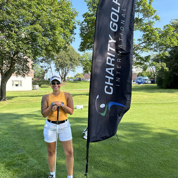 A person in a yellow tank top, white shorts, and a white cap stands smiling on a golf course during a 2023 Golf event, holding a golf club. Beside them is a tall flag with the text "CHARITY GOLF INTERNATIONAL." Trees and a building are visible in the background under the clear blue sky.