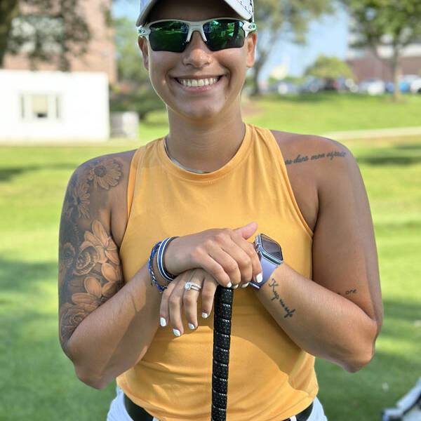 At a 2023 golf outing, a person wearing sunglasses, a white Titleist cap, a yellow sleeveless top, and white pants stands on the golf course smiling while holding a golf club. They have tattoos on one arm and are accessorized with bracelets and a smartwatch. Trees and buildings are visible in the background.