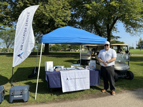 A man stands beside a booth with a blue canopy tent, which has a table displaying promotional materials for "Air Charter Service." A flag with the same logo is positioned to the left, while two golf carts are parked behind the booth on a grassy area under trees during the 2023 golf outing.