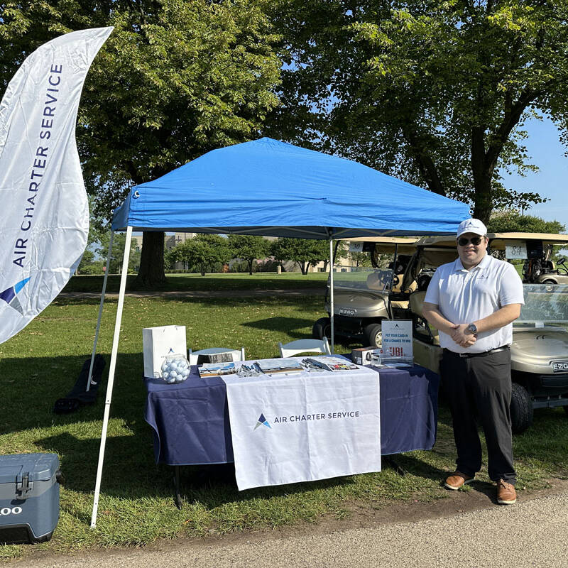 A man stands beside a booth with a blue canopy tent, which has a table displaying promotional materials for "Air Charter Service." A flag with the same logo is positioned to the left, while two golf carts are parked behind the booth on a grassy area under trees during the 2023 golf outing.