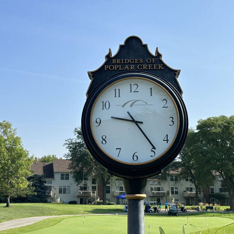 A black clock stands tall with the words "Bridges of Poplar Creek" at the top, reading approximately 10:12. In the background, trees, apartment buildings, and a person preparing to putt on a golf green set the scene for a perfect golf outing in 2023 under a clear blue sky.