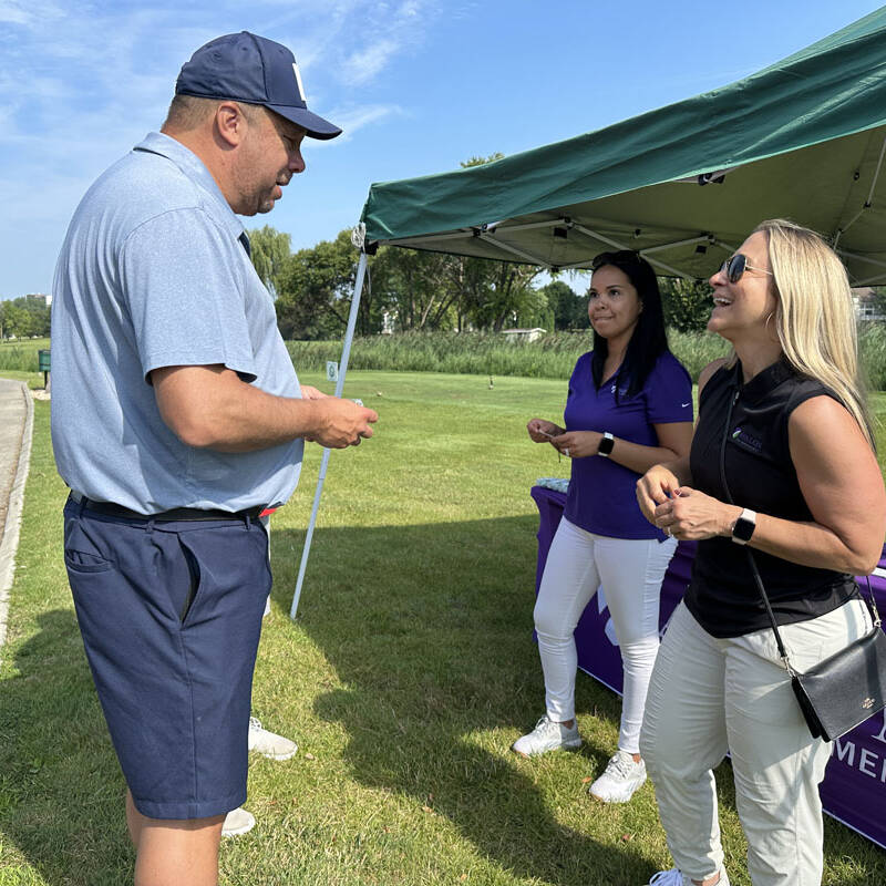 A man wearing a cap and shorts converses with two women under an outdoor canopy during a 2023 outing. One woman is dressed in a purple shirt and white pants, while the other wears a black sleeveless top and beige pants. A purple tablecloth with white text is on the table behind them, hinting at golf event arrangements.