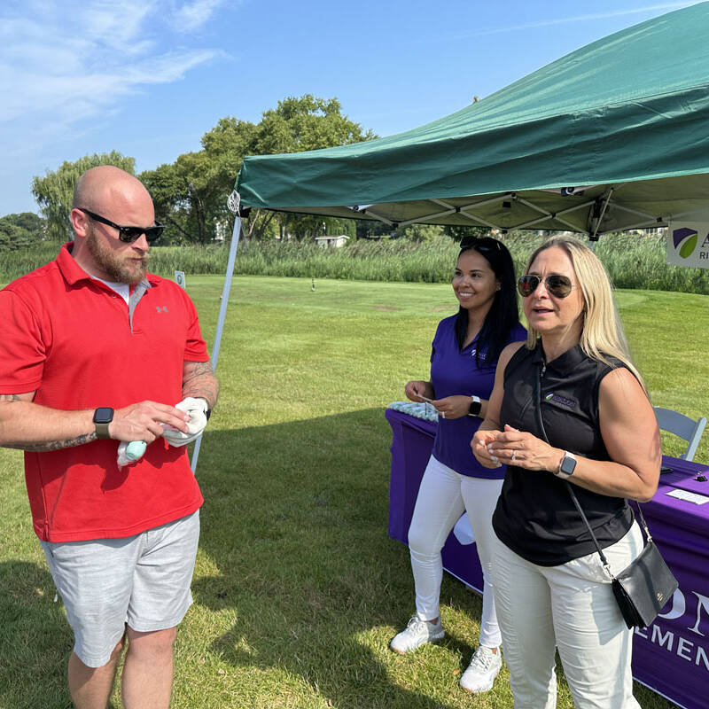 A man in a red shirt, wearing sunglasses and holding a cloth, stands next to a woman in a black sleeveless shirt and another woman in a purple shirt under a green canopy at the 2023 Avalon Risk Management outing. The background features grass, trees, and a clear sky.