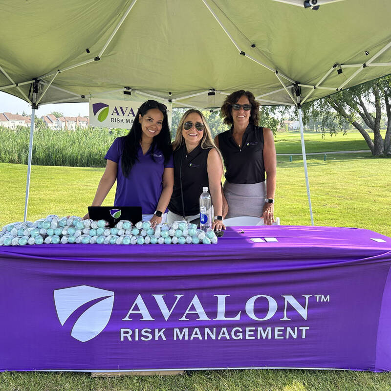 Three smiling women stand behind a purple table covered with golf balls and promotional items under a green canopy. The tablecloth and background banner feature the "AVALON RISK MANAGEMENT" logo. The setting appears to be a 2023 outdoor golf outing.