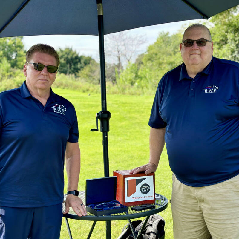 Two men wearing blue polo shirts with an "EWS" logo stand under a blue umbrella in a green outdoor setting. They are standing by a table holding a small box with the logo and text "Welcome to the team." One man is wearing sunglasses, and the other is not. It's 2023, and they're ready for the golf outing.