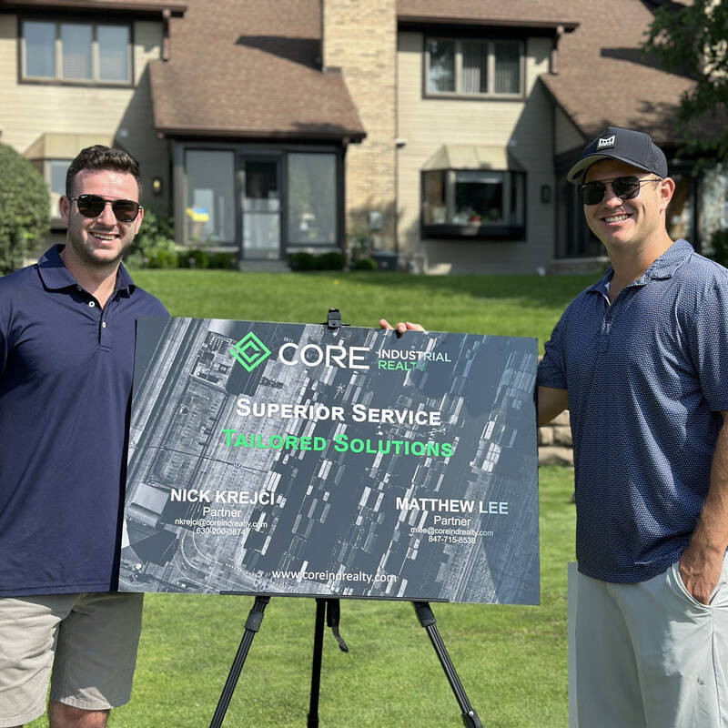 Two men wearing sunglasses and casual clothing stand on a lawn in front of a large sign for Core Industrial Realty. The sign reads "Superior Service, Tailored Solutions!" and includes the names Nick Krejci and Matthew Lee, who are identified as partners. Looks like a perfect day for a 2023 outing!