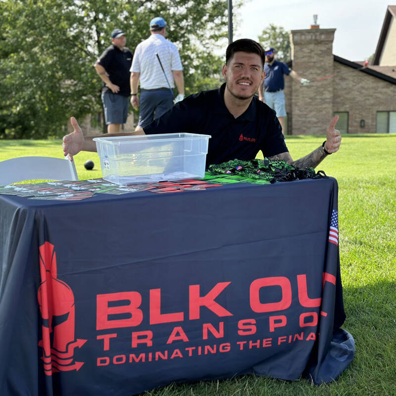 A person sits behind a table covered with a dark cloth that reads "BLK OUT TRANSPORTS: DOMINATING THE FINAL MILE." The table has pamphlets and a transparent bin on it. The person smiles and points outward, while in the background, a house, trees, and people at the 2023 Golf Event can be seen.