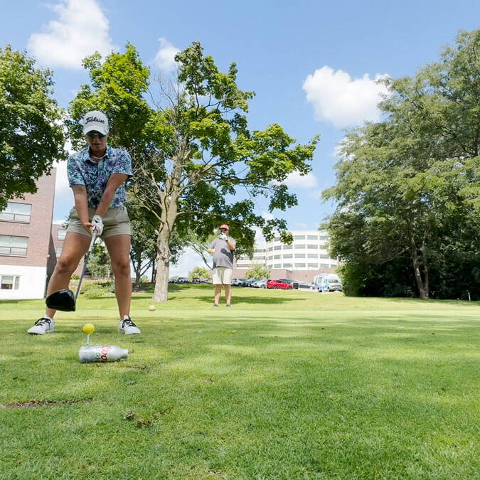 Two people are on a golf course during their 2024 outing. In the foreground, one person is preparing to tee off, focused on their shot. Another stands in the background, watching. The course is lush and green, surrounded by trees, with a building visible in the distance.