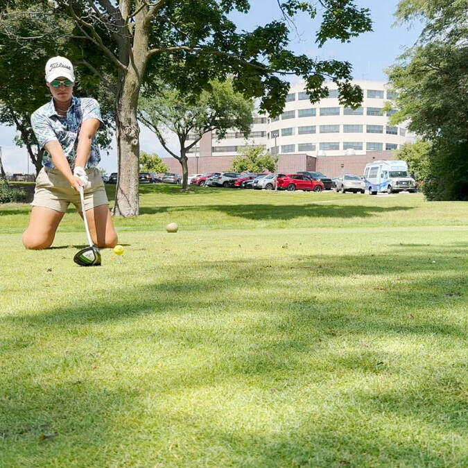 A golfer in shorts and a cap swings a club to hit a golf ball on a lush green course during the 2024 golf outing. Trees, a few parked cars, and buildings are visible in the background. The sunny weather casts clear shadows on the grass.