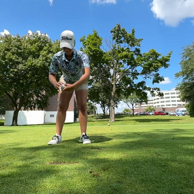 A person wearing a cap and sports attire prepares to tee off on a grassy golf course during a 2024 golf outing. Trees and a few buildings are visible in the background under a clear, blue sky. The golfer is focusing on the ball, appearing ready to make a shot.