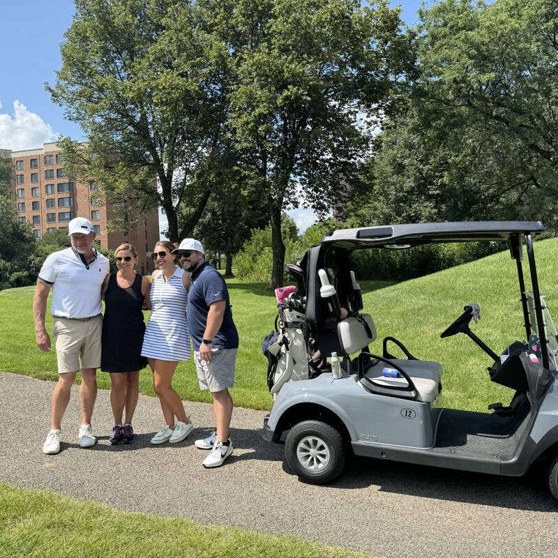 Four people stand together smiling on a sunny day next to a golf cart on a paved path during their 2024 outing. They are surrounded by lush green grass and trees, with a building visible in the background. Two women are in the middle between two men, clearly enjoying their time on the golf course.