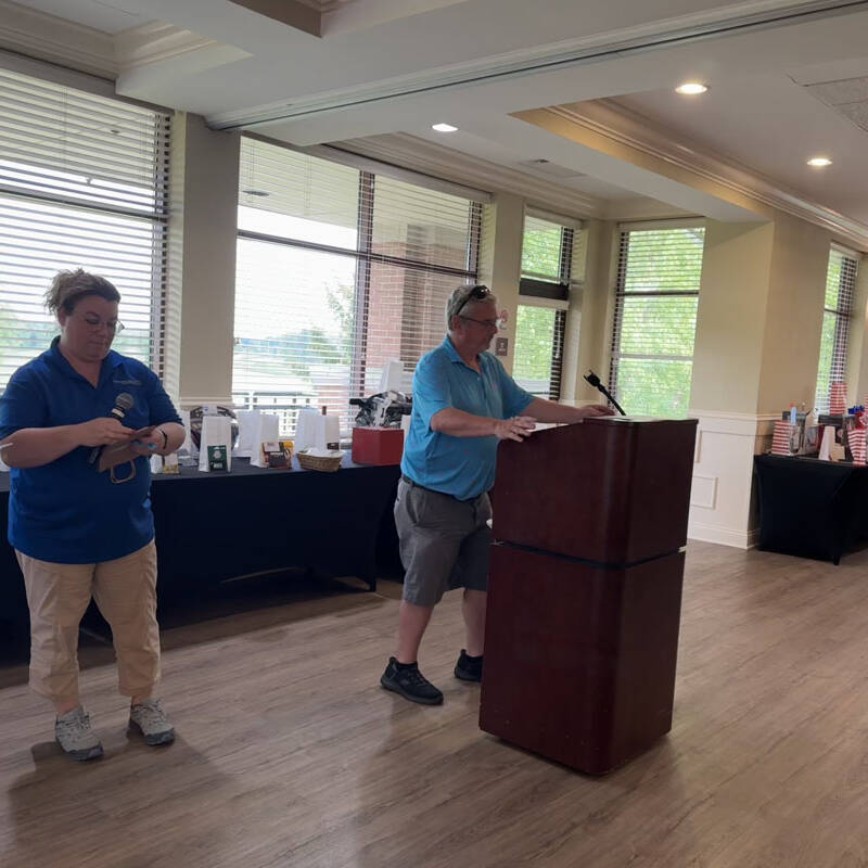 Three people are standing in a room with large windows. One person speaks at a podium, while a second person in the middle holds a microphone and looks at a piece of paper detailing the 2024 golf outing. A table laden with items lines the wall. Two of the people wear blue shirts.