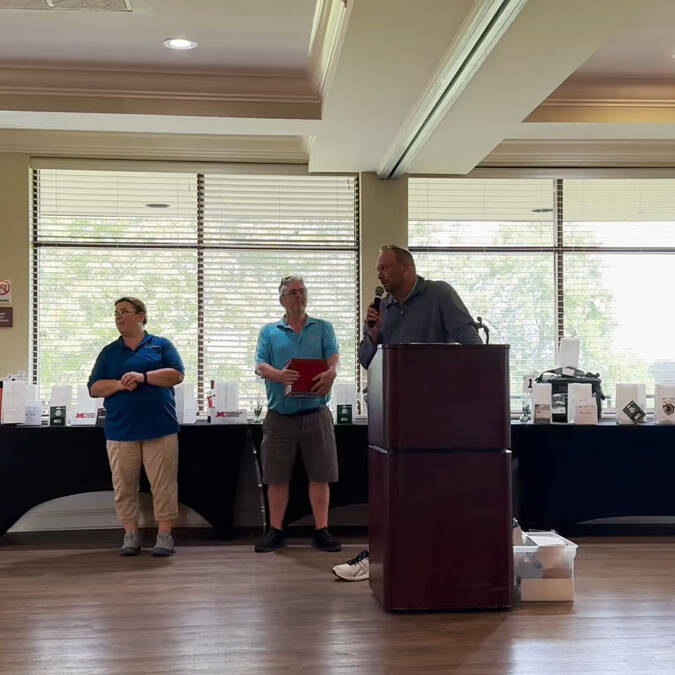 A man stands at a podium speaking into a microphone in a room with large windows. To his left, another man holding a red folder and a woman in a blue shirt are standing. In the background, tables display various items for the 2024 Golf Outing.