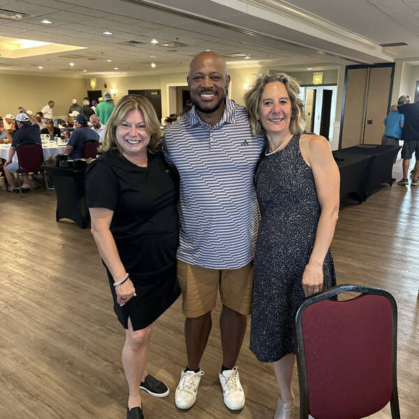 Three people stand smiling in a banquet hall with several tables and guests in the background, celebrating a successful 2024 Golf outing. The person on the left wears a black dress, the person in the middle wears a striped shirt and shorts, and the person on the right wears a dark dress.