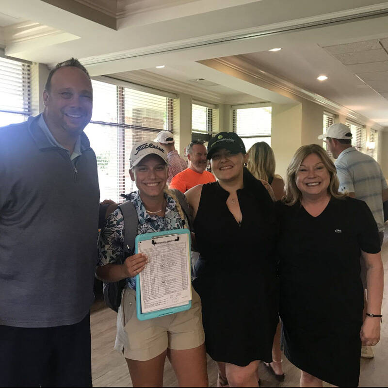 Four people are posing for a photo indoors at a golf event. A woman in the center is holding a clipboard, and the other three are standing around her, smiling. There are additional people in the background socializing.