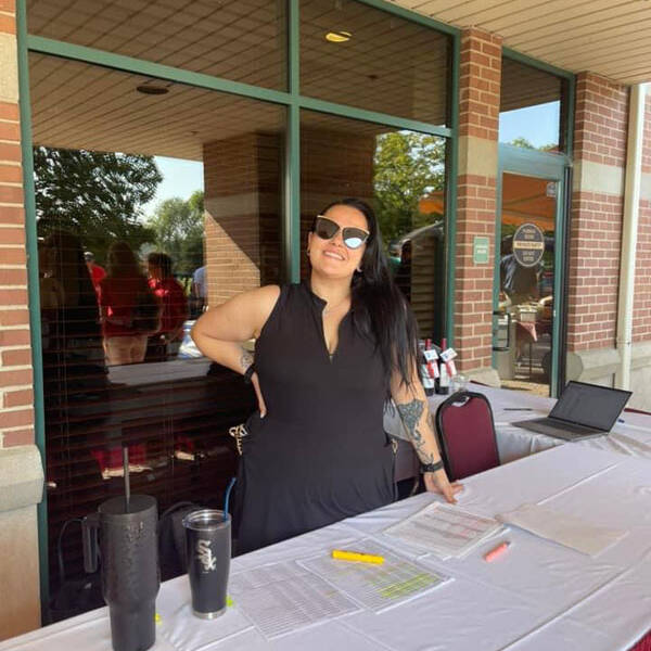 A person with long dark hair, wearing sunglasses and a black sleeveless dress, stands behind a table covered with a white cloth. The table has a laptop, several papers, and a few drink cups. They are outdoors in a shaded area near a brick building, managing registrations for the 2024 Golf Outing.