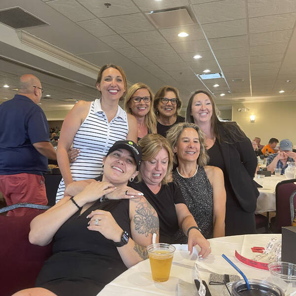 A group of seven women are gathered in a room with white ceiling tiles and recessed lighting. Four are seated at a round table with drinks, and three stand behind them. Most are smiling or laughing, creating a lively and joyful atmosphere as they plan their 2024 Golf Outing.