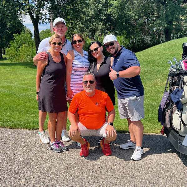 A group of six people are posing together outdoors on a sunny day during the 2024 Golf Outing. They are on a golf course, with golf carts and trees visible in the background. Two of them are wearing golf attire, and one person, dressed in orange, is crouching in front while the others stand behind.