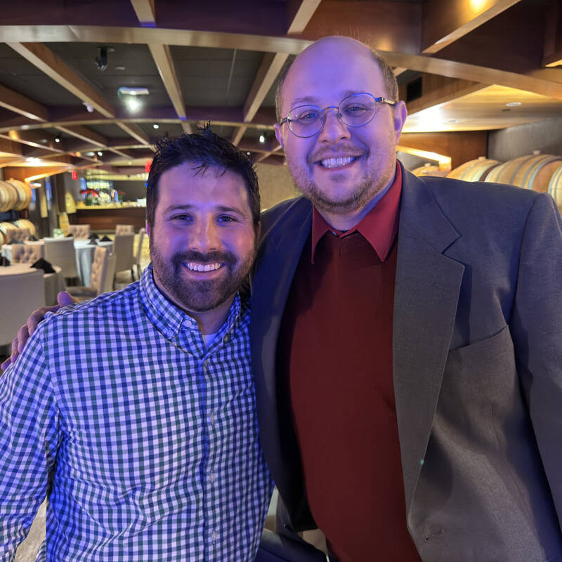 Two men are smiling as they pose for a holiday photo in a restaurant. The man on the left is wearing a checkered shirt, while the man on the right sports glasses, a red sweater, and a gray blazer. The background features dining tables and large wooden barrels, adding to the festive 2023 vibes.