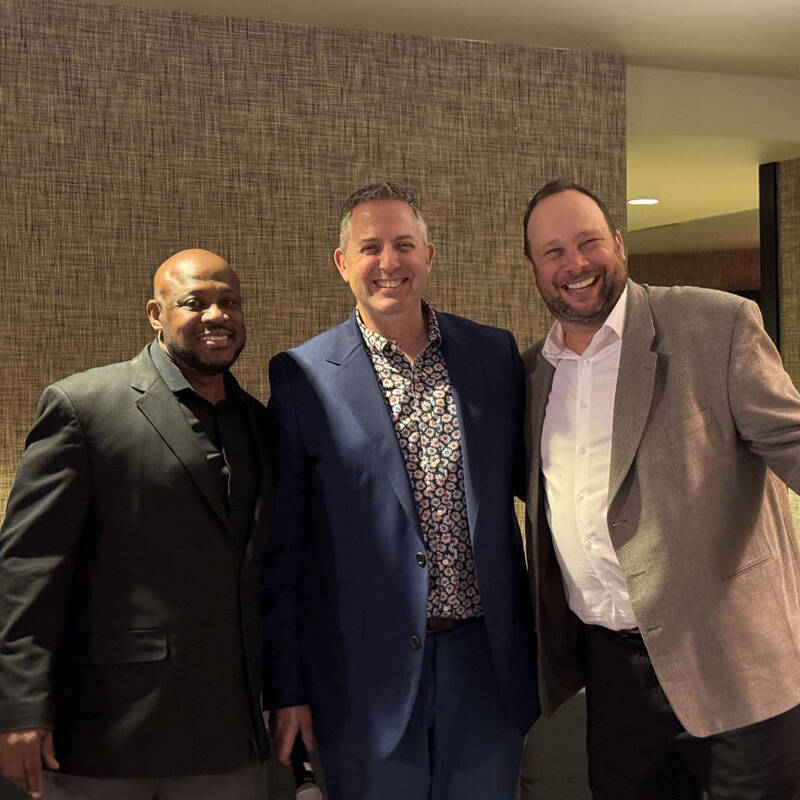 Three men are standing together and smiling at a holiday party in 2023. They are dressed in formal attire; one in a black jacket, the second in a blue suit, and the third in a grey suit. The background features a textured wall with subtle decorations.