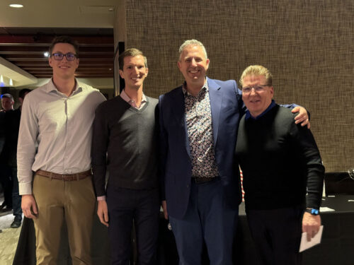 Four men standing together and smiling at the camera in a well-lit indoor setting. The man on the far left is wearing glasses, a light-colored shirt, and khaki pants. The men in the center and on the right are wearing sweaters, while the man second from the right sports a suit jacket, ready for a 2023 holiday party.