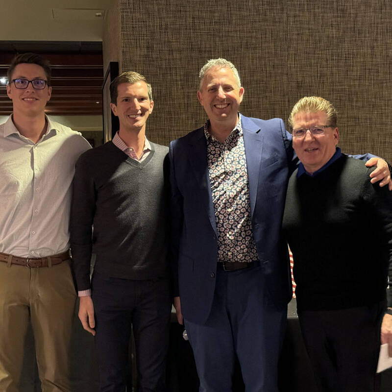 Four men standing together and smiling at the camera in a well-lit indoor setting. The man on the far left is wearing glasses, a light-colored shirt, and khaki pants. The men in the center and on the right are wearing sweaters, while the man second from the right sports a suit jacket, ready for a 2023 holiday party.