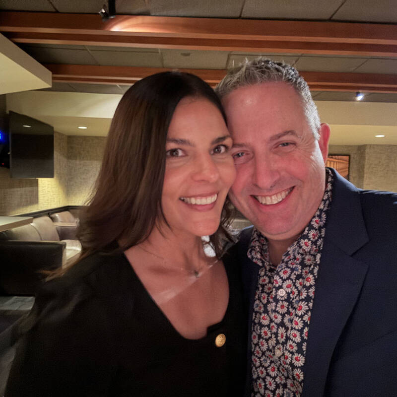 A woman with long dark hair and a man with short grey hair, both smiling and standing close together at a holiday party in 2023. The warmly lit indoor setting features modern decor. The woman is wearing a black top, while the man sports a dark blazer over a floral shirt.