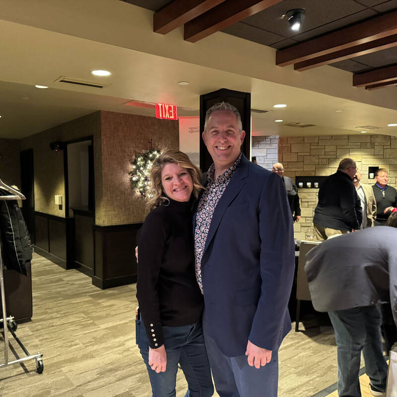 A woman and a man stand close together, smiling at the camera in a warmly lit indoor setting. Other people are in the background engaging in conversation and activities at the 2023 holiday party. The room features modern decor with a stone wall and coat rack.