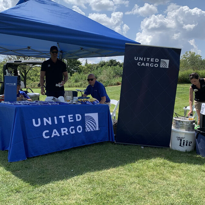 A United Cargo promotional tent is set up on a grassy area under a sunny sky during the 2024 Golf Outing. Two men stand behind a table covered with a blue United Cargo tablecloth. To the right, a woman stands near a cooler filled with drinks. A branded banner is displayed prominently.