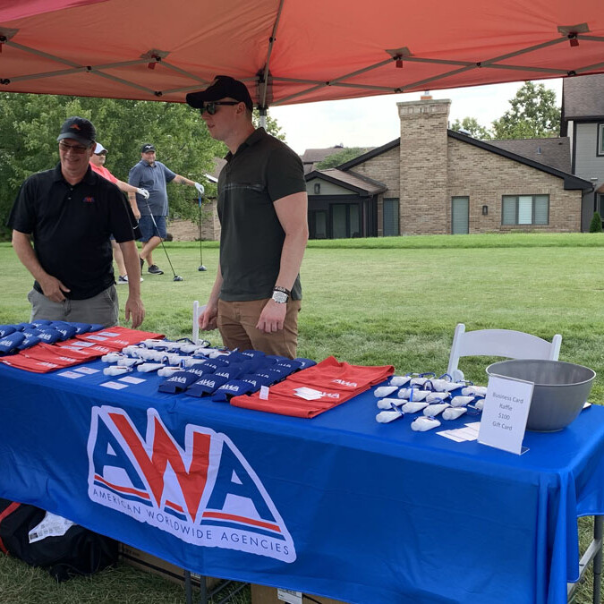 At an outdoor event in 2024, a group of people is gathered around a booth covered by a red tent. The table, adorned with a blue tablecloth displaying "Golf Tee Holders," features two men behind it and one person engaged in conversation. This lively outing highlights the excitement for all things golf-related.