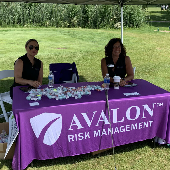 Two women sit at a purple table under a canopy at an outdoor event. The table, draped with a branded Avalon Risk Management tablecloth, is covered with small items or treats. Set against the backdrop of tall vegetation and grassy terrain, this scene is part of the 2024 Golf Outing.