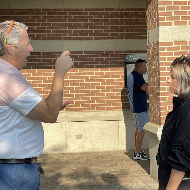 A man and a woman are engaged in conversation outside a brick building, possibly discussing their recent golf outing. The man gestures with his hands while the woman listens attentively. Another person stands in the background, partially in shadow. Both people in the foreground have light-colored hair.