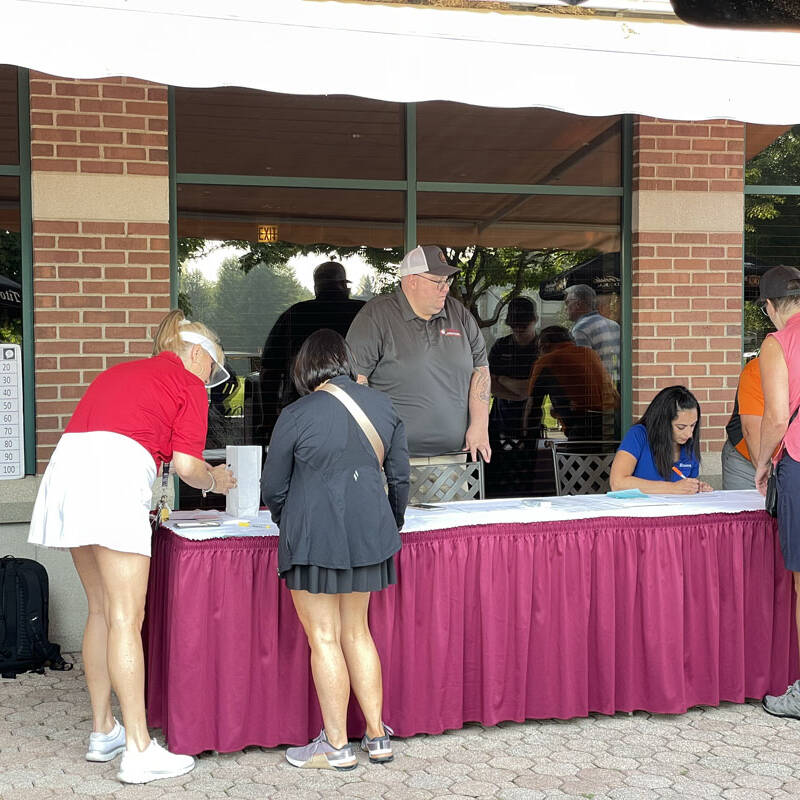 People are gathered at an outdoor registration table covered in a red tablecloth. Two individuals are filling out forms for the 2023 golf outing, while others are providing assistance. A brick building with windows is in the background, and a schedule is posted on the wall.