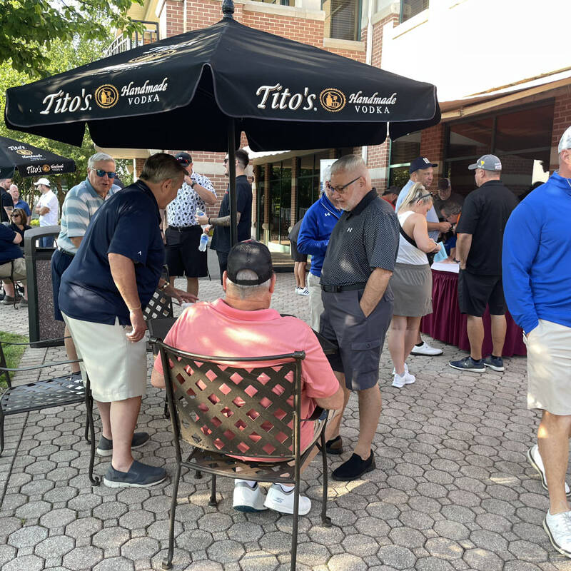 A group of people socializing on a patio under Tito's Vodka umbrellas during a 2023 outing. Some are seated while others stand and talk. The building in the background has large windows. A few attendees are near a table set up with informational brochures. It's a bright day perfect for golf discussions.