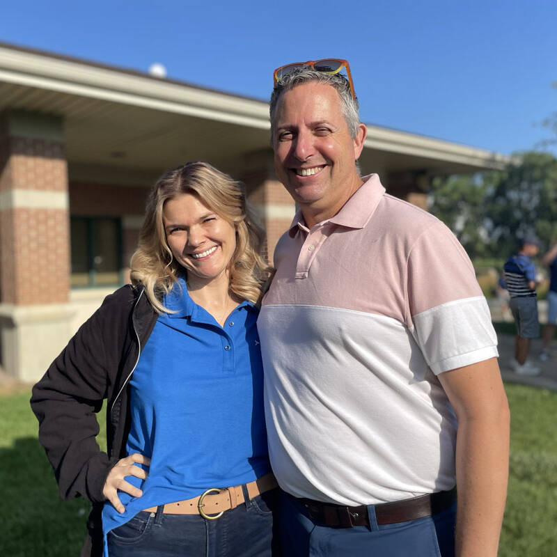 A woman in a blue shirt and a man in a pink and white shirt stand side by side outside a building on a sunny day during their 2023 outing. Both are smiling at the camera with other people visible in the background.