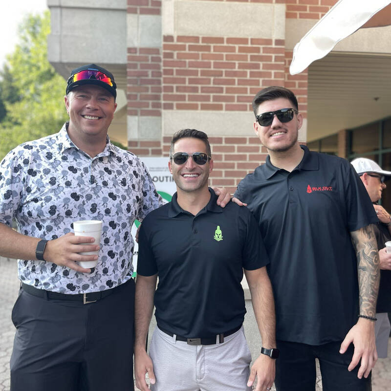 Three men standing together outside a brick building, smiling at the camera during their 2023 outing. The man on the left is wearing a patterned shirt, a baseball cap, and holding a cup. The man in the middle is in a black polo shirt, while the man on the right sports sunglasses and a black shirt.