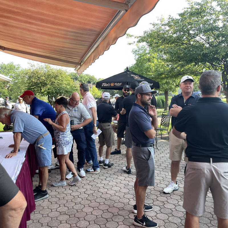 A group of people standing in line and interacting outside a building at a 2023 outing. Some are near a table with a maroon tablecloth, while others are conversing next to a shaded area with trees and an umbrella in the background, enjoying the social event or perhaps discussing their recent round of golf.